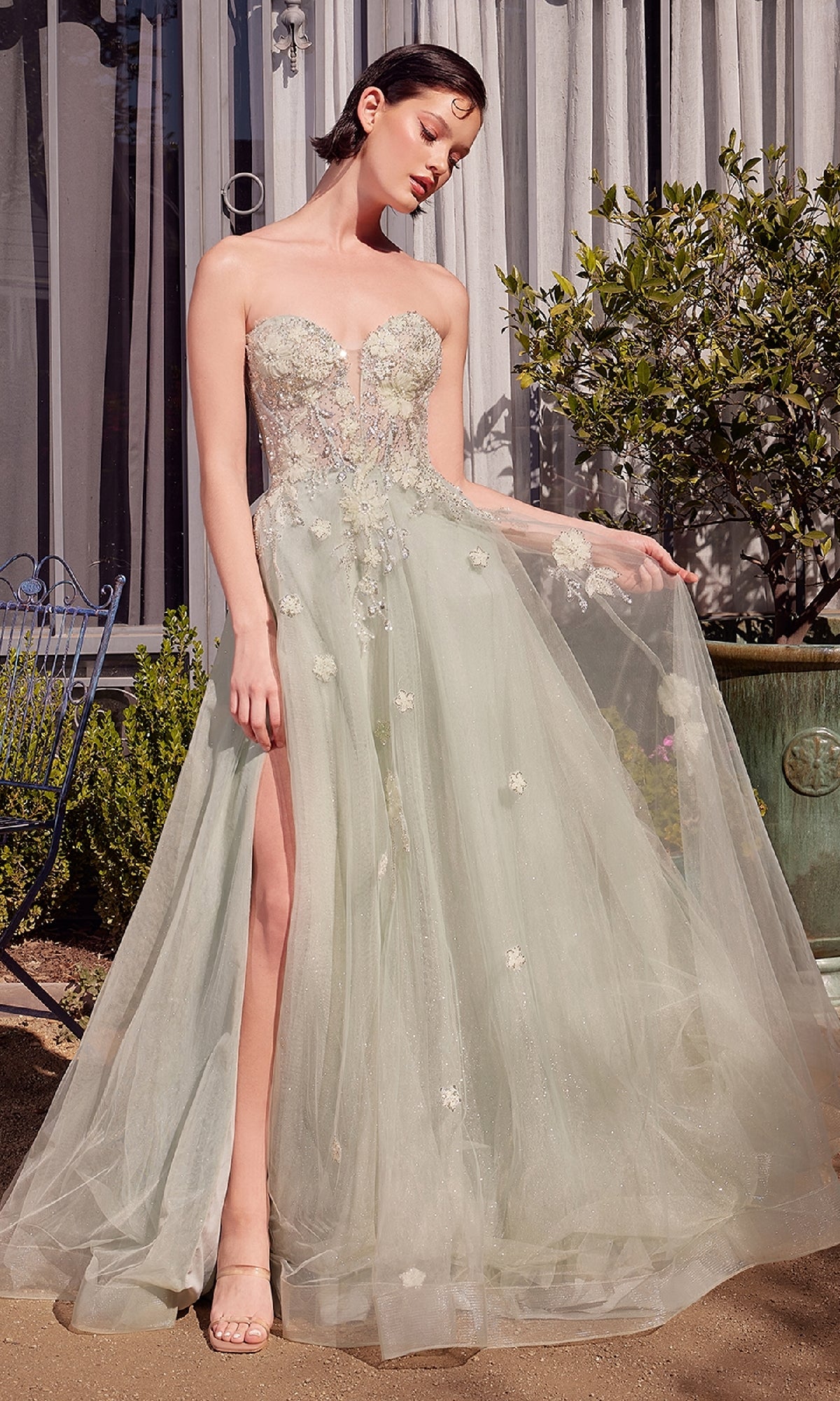 A woman stands outdoors in the Andrea & Leo A1418 Sage Green Prom Ball Gown, a strapless corset dress with floral embellishments and a high slit, holding part of the skirt out to the side.
